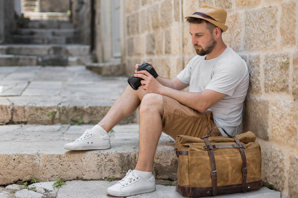 Man sitting on steps with a camera beside a waxed canvas duffle bag in a sunlit urban setting.