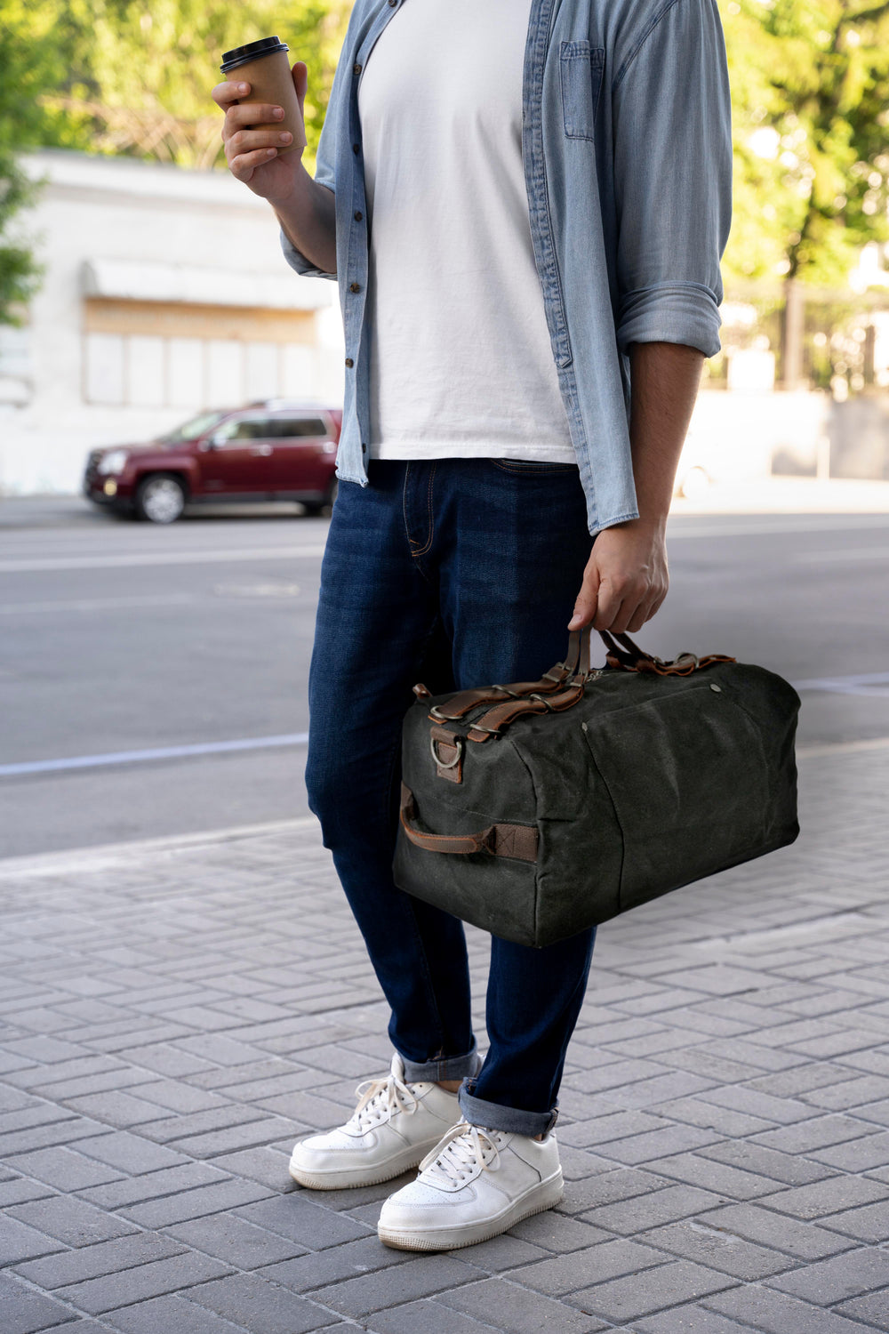 Man holding a sports duffle bag while enjoying coffee on a city street, dressed casually with sneakers.