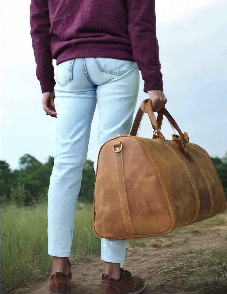 Man holding a brown leather holdall bag while walking on a path, showcasing style and functionality.