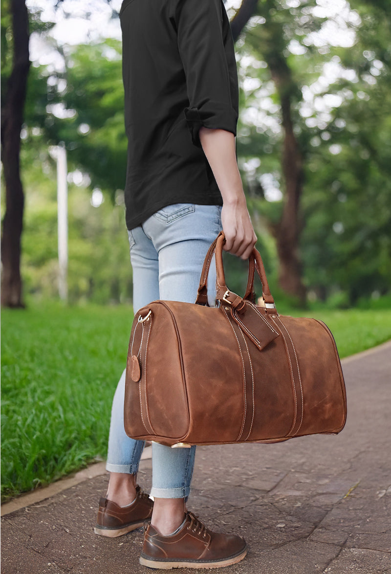 A person walking in a park carrying a stylish leather holdall bag, showcasing its rich brown color and elegant design.