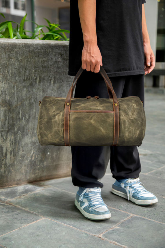 Person holding a military duffle bag with leather accents, dressed in casual attire and sneakers, outdoor setting.