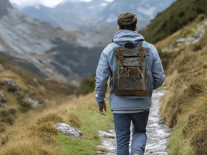 Man walking on a trail in the mountains, wearing a backpack and a hoodie, surrounded by nature.