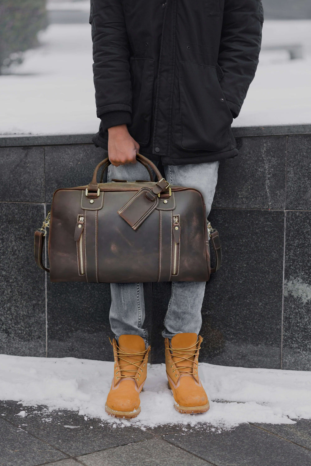 Man holding a stylish leather overnight bag in winter attire against a snowy backdrop.