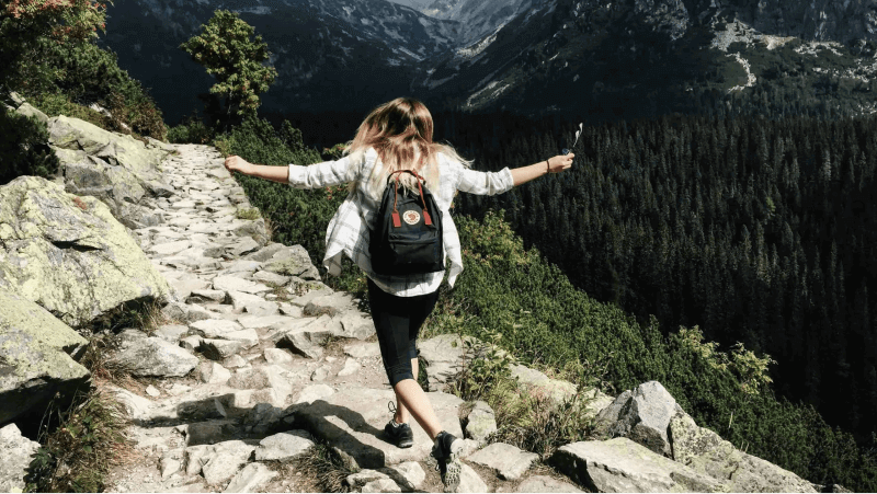 A hiker joyfully walking on a rocky trail surrounded by lush forests and mountains.