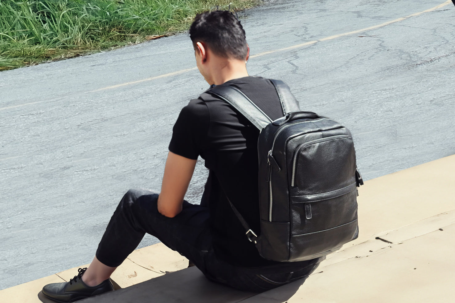 Man seated wearing a stylish black leather bookbag, showcasing modern design and functionality in a scenic outdoor setting.