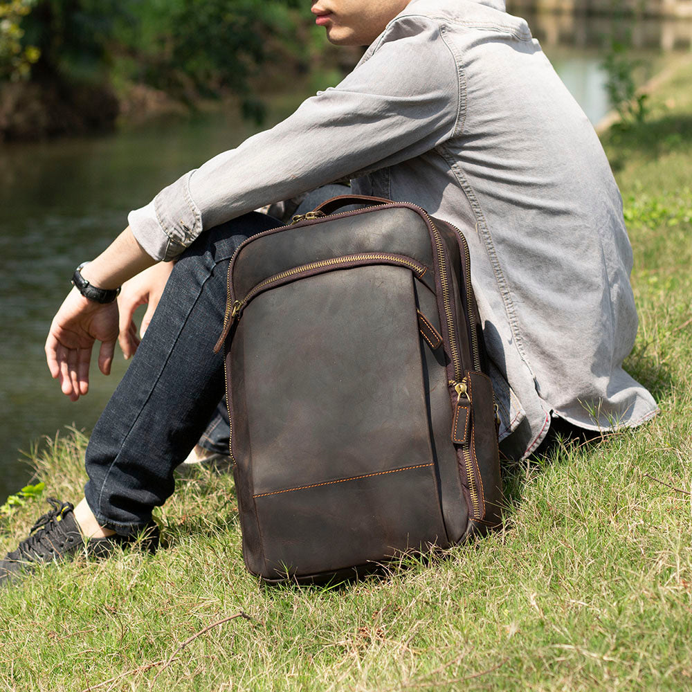 Leather laptop rucksack named Loch Ness, casually styled, placed beside a person sitting on grass by a river.