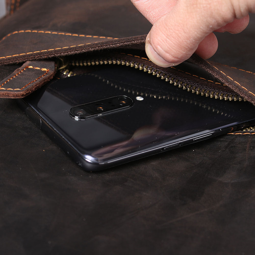 Close-up of a hand reaching into a leather bag, revealing a smartphone in a zipper compartment.