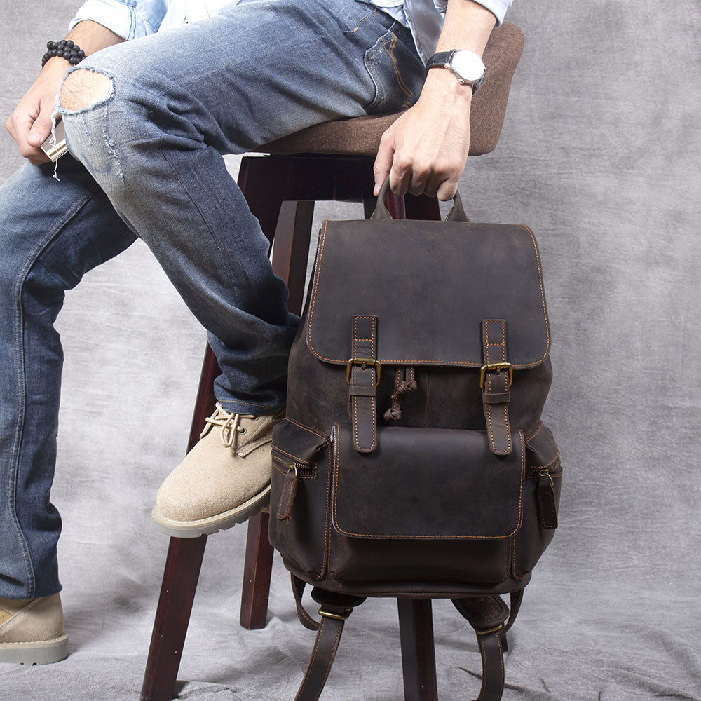 Stylish leather drawstring backpack in brown, displayed on a stool with a seated individual wearing casual attire.