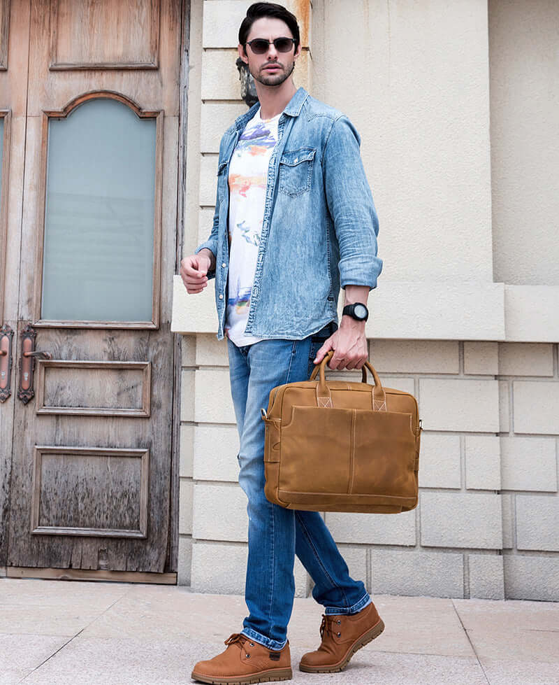 Stylish man in denim jacket holding a vintage leather messenger bag, walking near a wooden door.